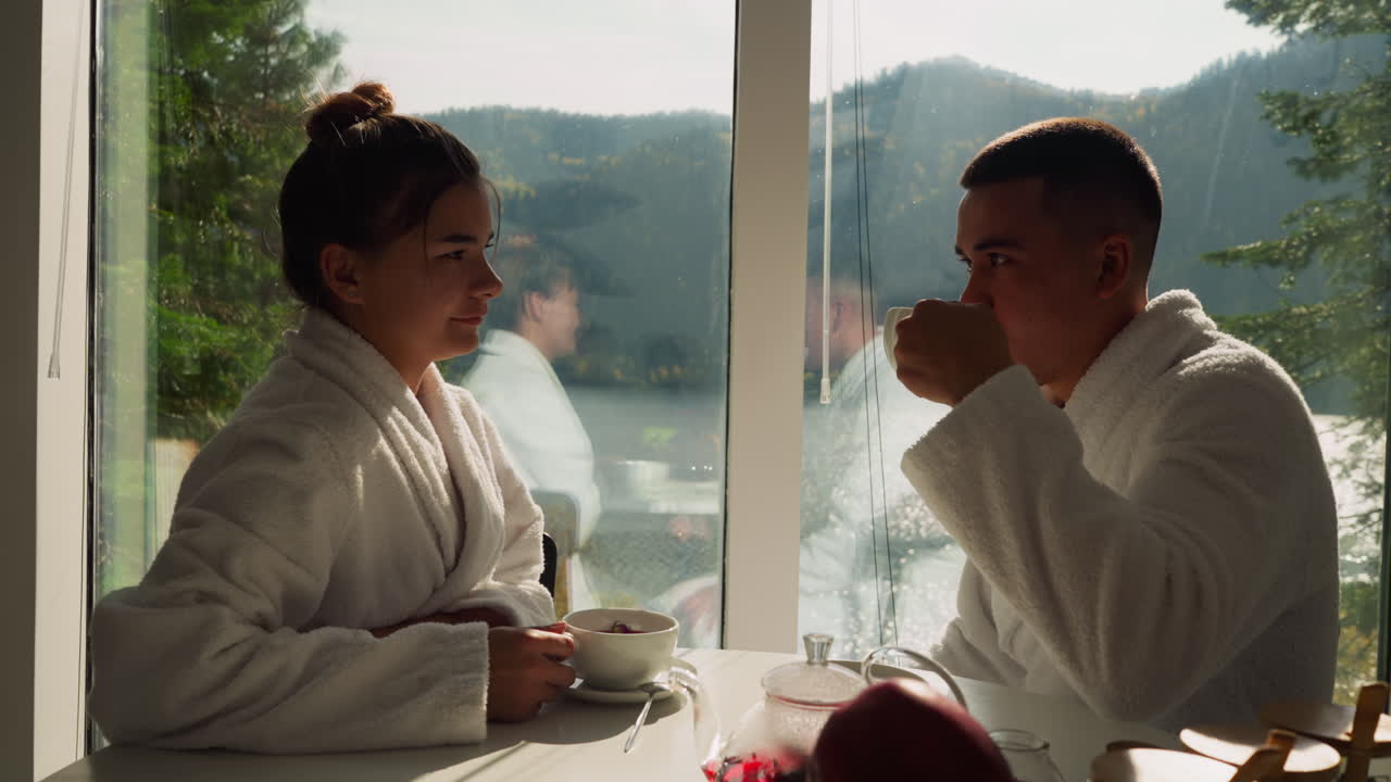 신혼부부는 부에서 창문 에 앉아 차를 마시고, 목욕복을 입은 젊은 부부는 아침 아침 식사에서 뜨거운 음료를 즐긴다.