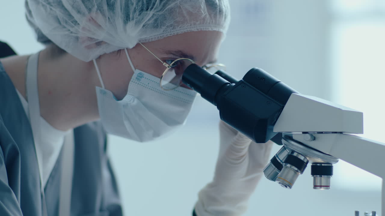 보호 의복 을 입은 여성 과학자 가 실험실 에서 현미경 을 사용 하고 있다