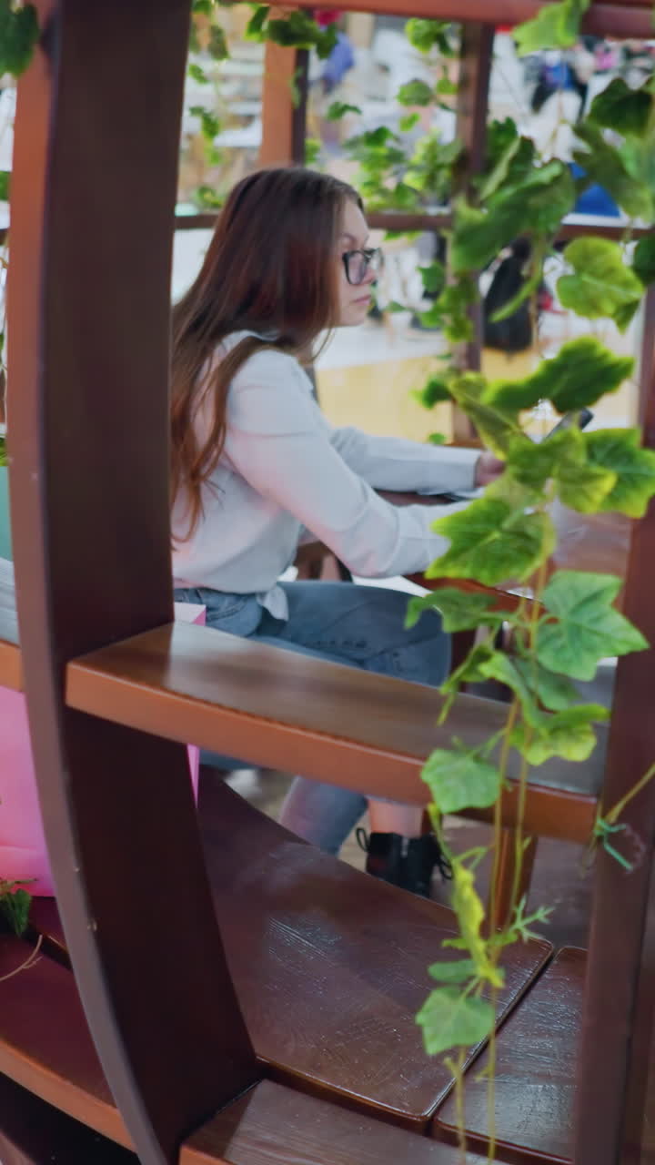 장식 된 나무 좌석에 앉아있는 멋진 젊은 여성, 전화를 들고, 그녀의 에 쇼핑 가방, 쇼핑몰의 바쁜 레스토랑 배경에서 움직이는 사람들과