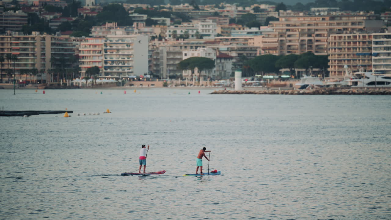 Two people paddleboarding together near a coastal city with modern buildings