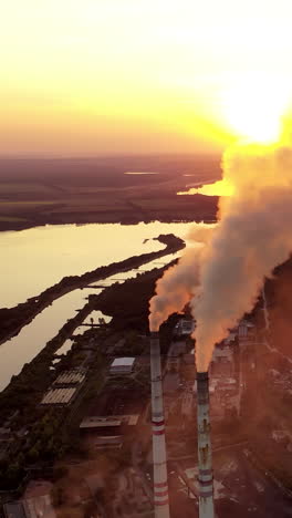 pipes with smoke on the natural green fields background. clouds of smoke coming from the chimneys of the plant with yellow lighting of setting sun. aerial view. vertical video