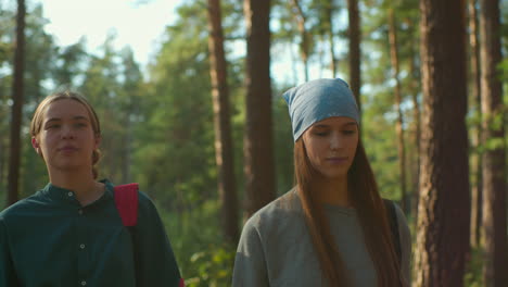 two friends walking through a sunlit forest, one wearing a blue headscarf and the other with a red backpack, warm smiles and gentle expressions as sunlight filters through tall trees