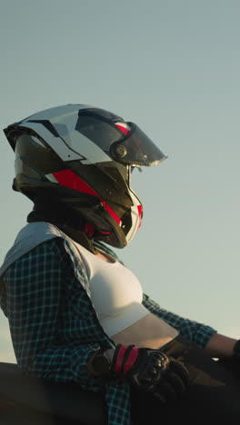 a female biker is seen resting on her red power bike, wearing a helmet and gloves, as she looks forward into the open field