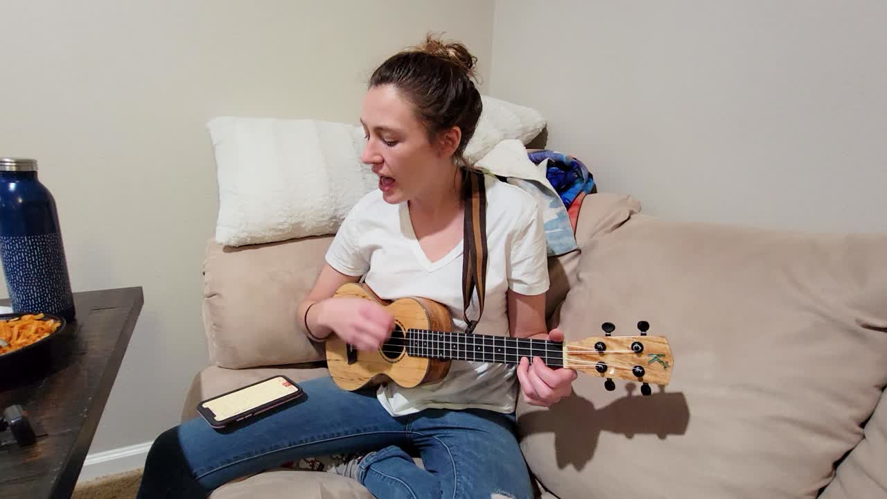 Donna canta mentre suona l'ukulele su un divano al chiuso, eseguendo musica a casa con un'impostazione acustica informale, testi sullo smartphone nelle vicinanze e una scena di vita intima e rilassata.
