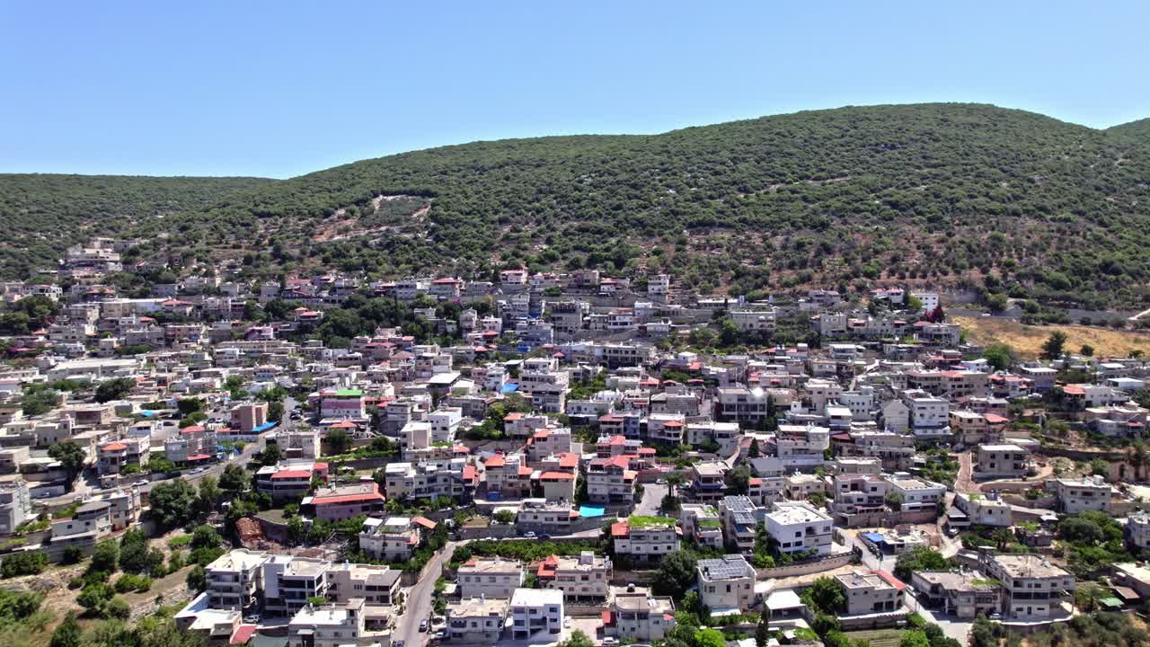 Aerial view of the Druze town of Peki'in in northern Israel, surrounded by lush green mountains under a clear summer sky.