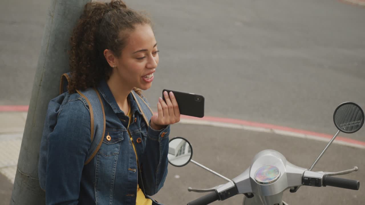 Happy biracial woman in city, sitting on motorbike using smartphone