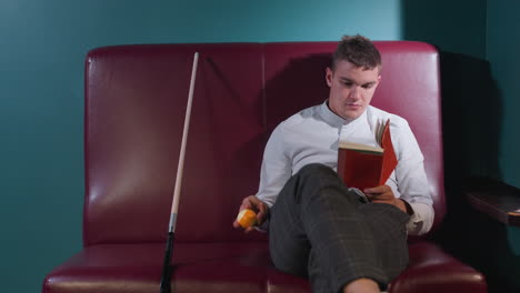 chap in white shirt and plaid pants sits on red sofa in billiard room, reading book while rolling orange billiard ball in hand. cue stick leans beside him