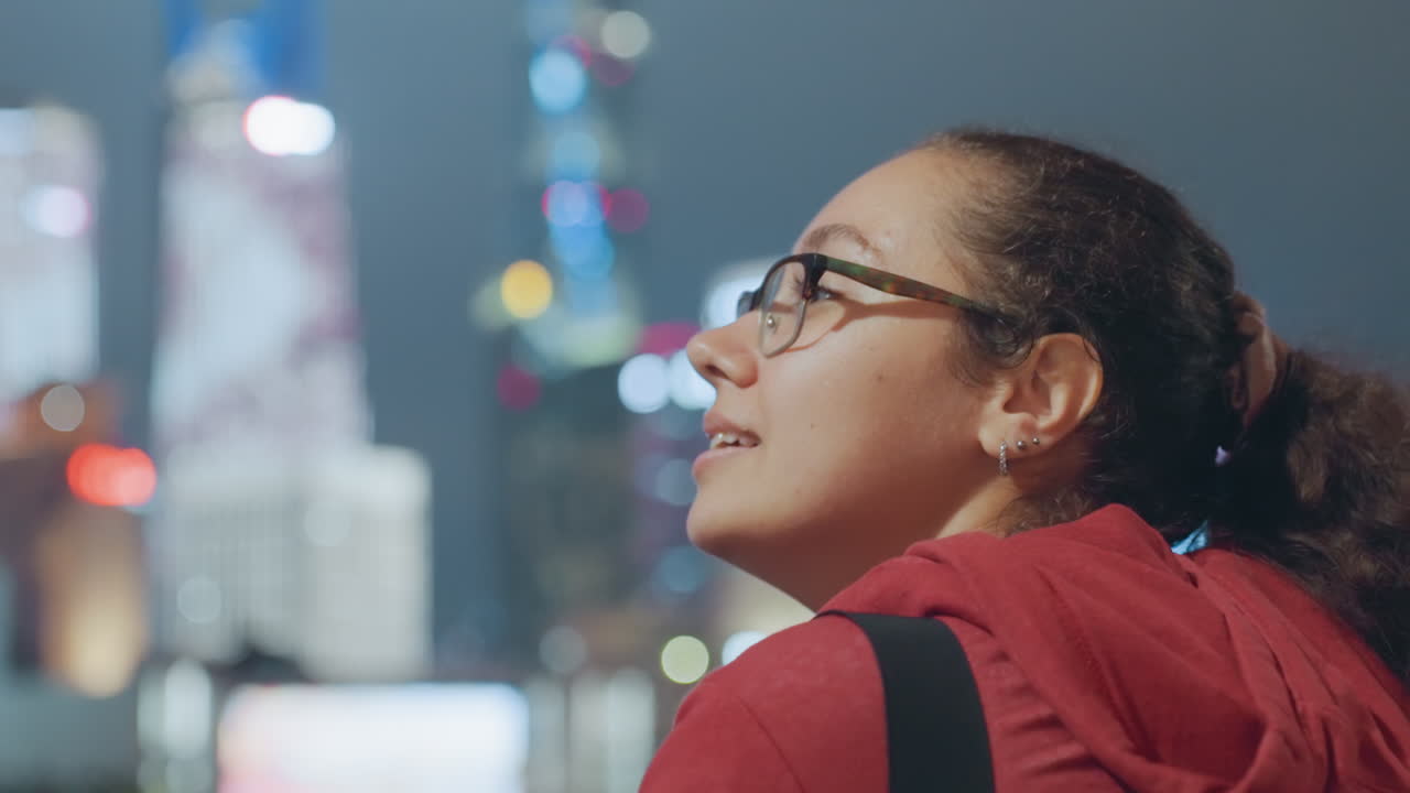 Young woman in red hoodie skyline, glasses and ponytail, gazing across illuminated waterfront with blurred city lights contemplative atmosphere with slowmotion profile shots, frames suggest student
