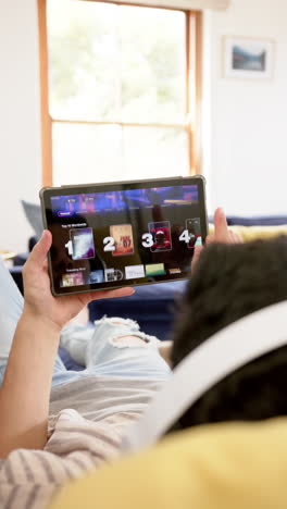 vertical video of biracial man in headphones lying on couch using tablet, slow motion