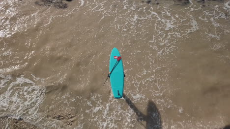 vista de pájaro del hombre caminando a lo largo de guijarros hacia el mar con una tabla de remo equilibrada en su cabeza