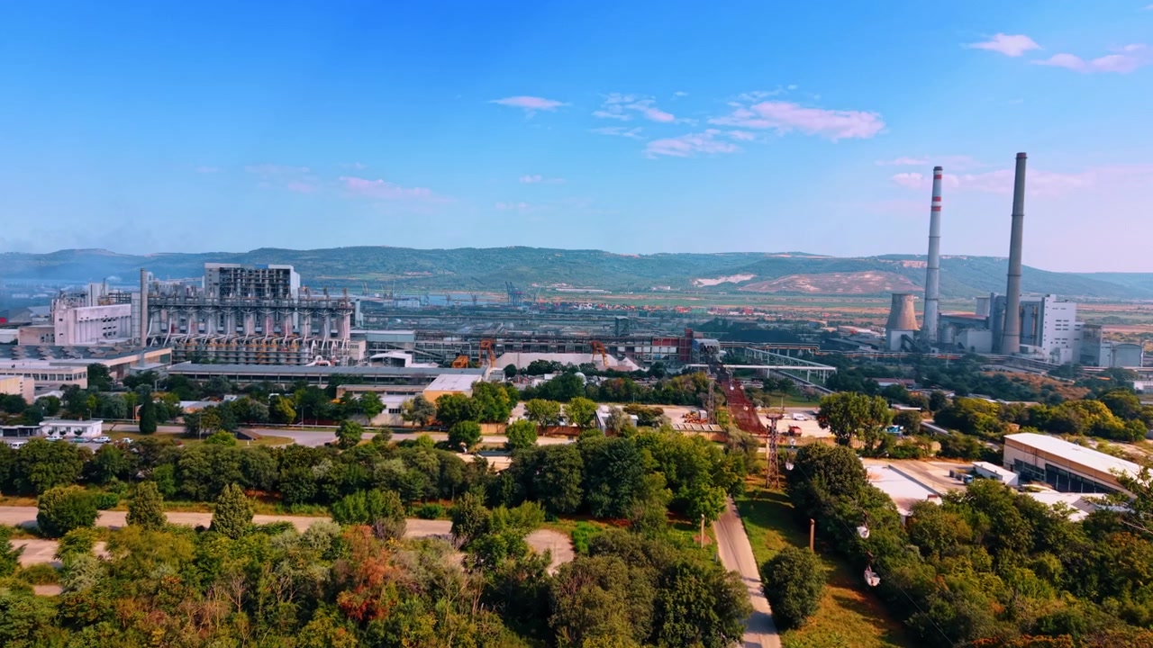 Fjernt luftfoto af et industrianlæg omgivet af skov. Tæt grønt løv dækker forgrunden, der fører mod fabriksbygninger og høje tårne.