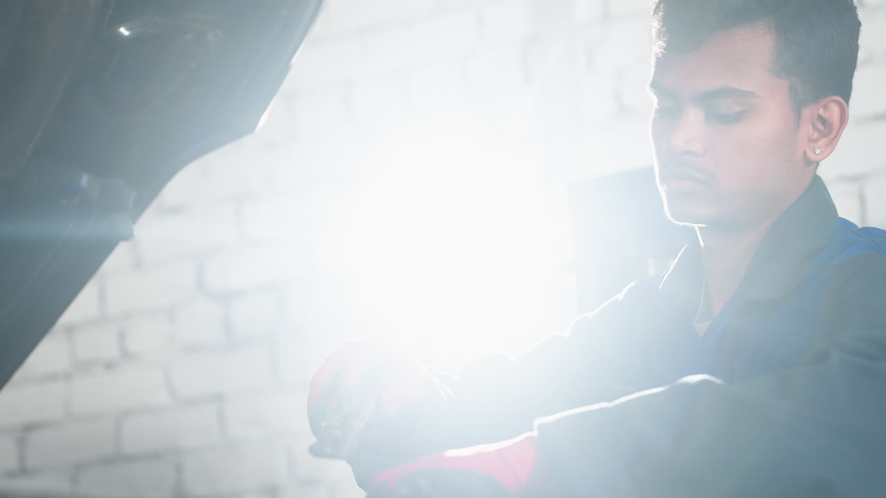 ingeniero en uniforme azul cambia de herramienta mientras trabaja en el motor del coche en un garaje bien iluminado, una luz brillante brilla en el fondo, añadiendo énfasis a la tarea enfocada a la mano