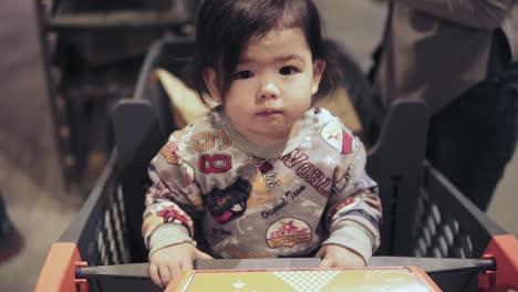 Cute-asian-toddler-boy-is-sitting-in-a-shopping-cart-while-his-mom-is-picking-out-groceries-at-supermarket.