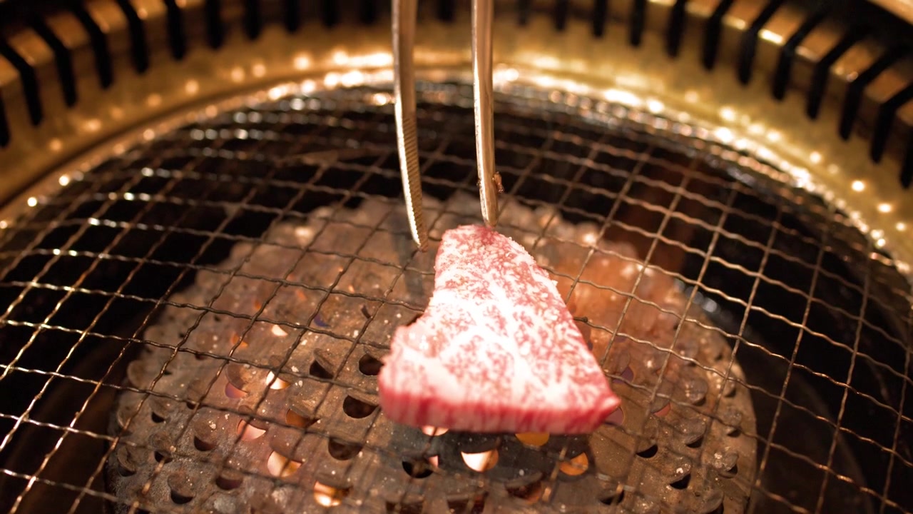 Primo piano di carne di manzo marmorizzata che cuoce su fiamme di carbone in un ambiente da ristorante giapponese.