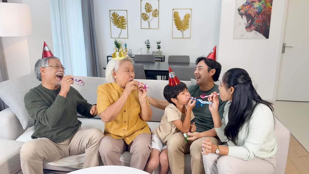 Four generations celebrate a birthday with party hats and laughter in a bright living room