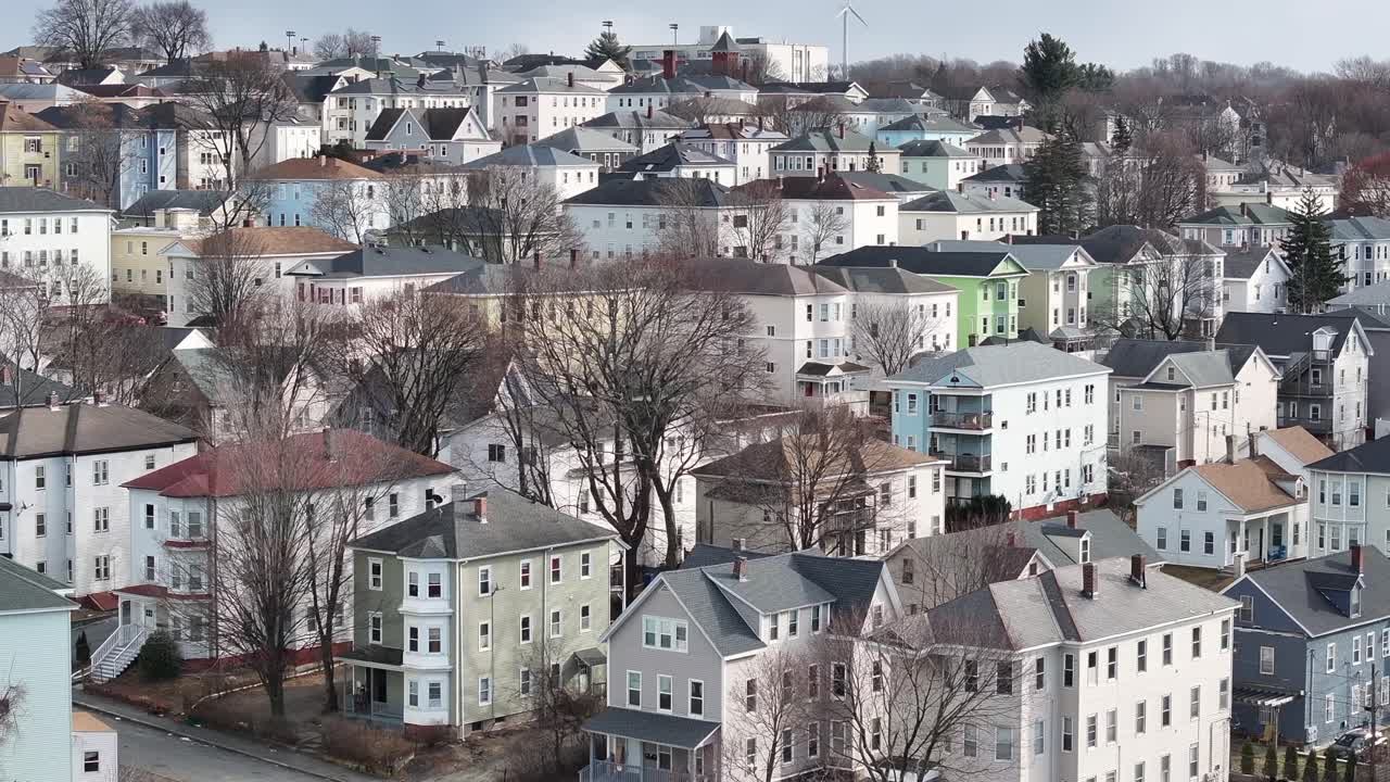 Triple-decker homes fill Worcester, Massachusetts hillside. Painted exteriors span a bustling residential block. Over rooftops, aerial.