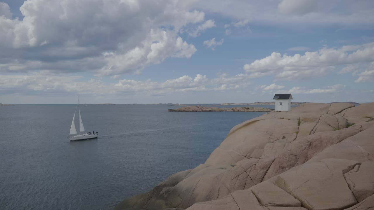маленький дом на побережье швеции на скале с парусной лодкой, проходящей мимо