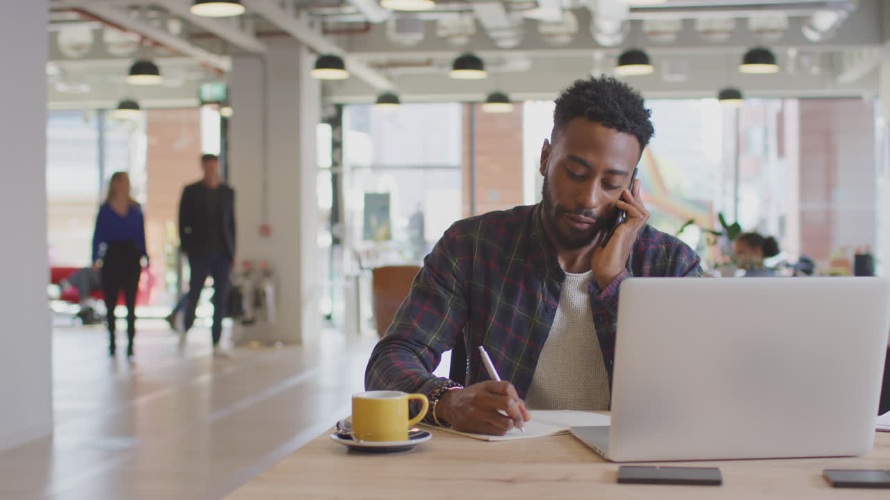 Businessman Sitting At Desk On Phone Call In Modern Open Plan Office With Colleagues In Background