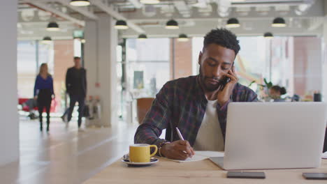 businessman sitting at desk on phone call in modern open plan office with colleagues in background