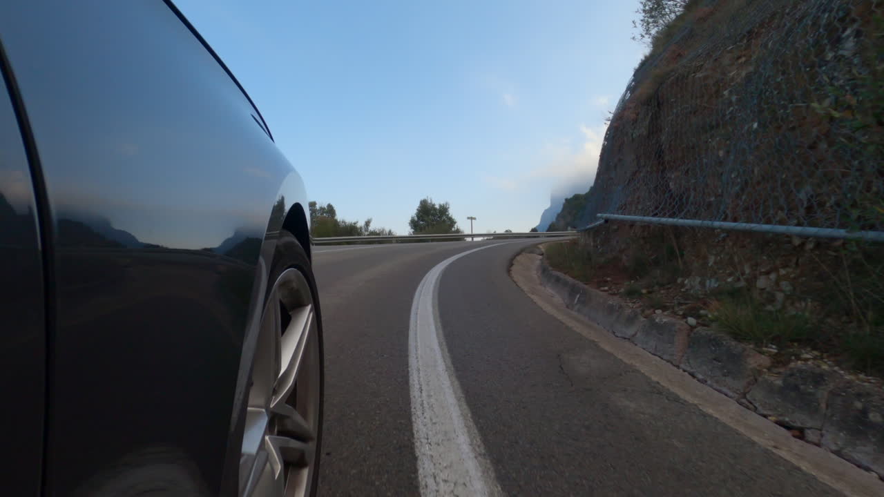 Visuale dal lato di un'auto, con angolazione bassa (POV), che percorre una tortuosa strada di montagna.