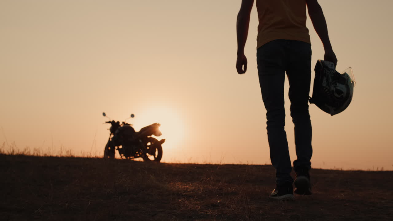 Un uomo con un casco in mano cammina verso la sua moto al tramonto