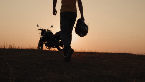 Ein-Mann-Mit-Einem-Helm-In-Der-Hand-Geht-Bei-Sonnenuntergang-Zu-Seinem-Motorrad