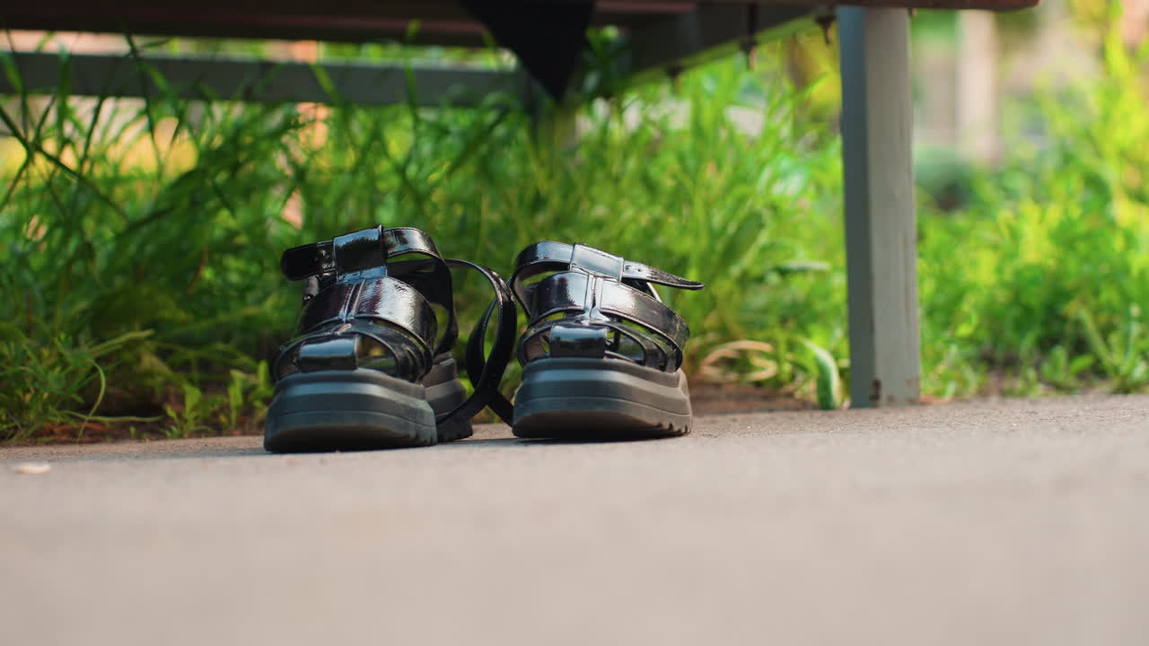 공원 벤치 아래 나란히 놓인 신발 클로즈업 사진입니다. 나무 벤치 옆 콘크리트 바닥에 검은색 가죽 신발이 나란히 놓여 있고, 뒤로는 무성한 녹색 잎이 배경으로 펼쳐져 있으며, 부드러운 오후 햇살이 비치는 모습에서 동반자적인 분위기가 느껴져요.