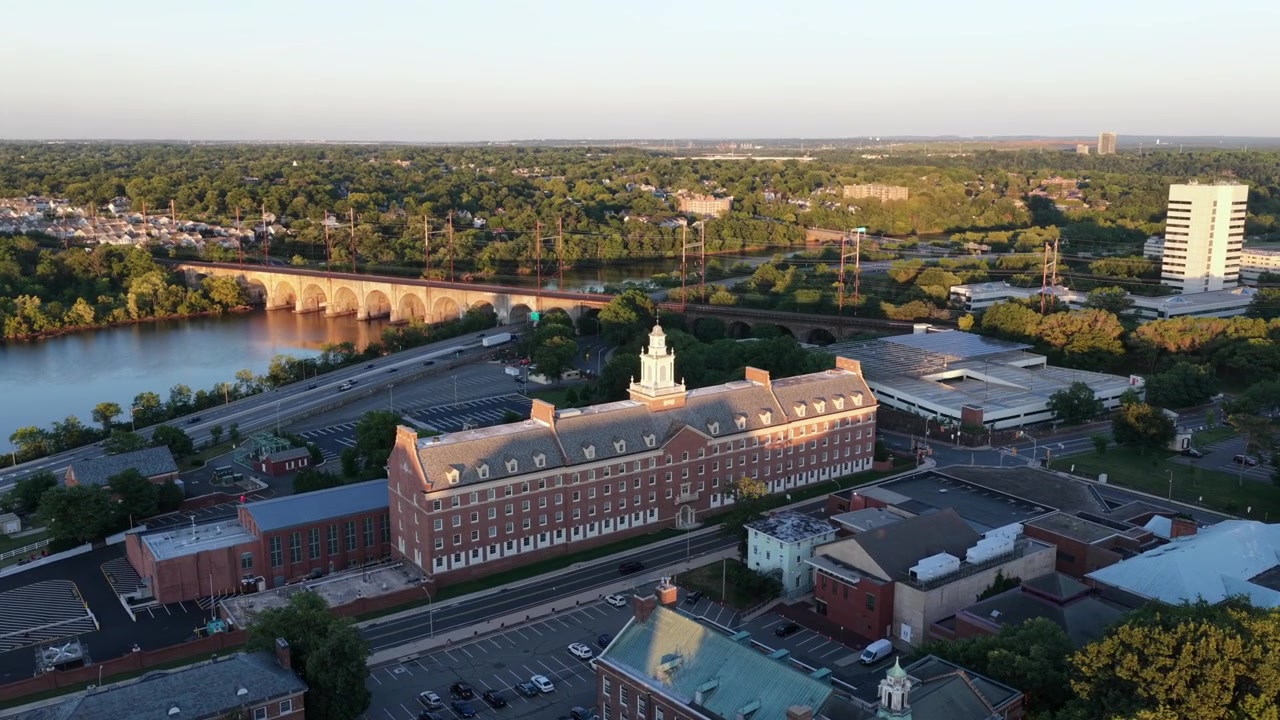 Storico edificio della Rutgers University si erge sopra il campus lungo il fiume a New Brunswick, New Jersey, al tramonto, con un ponte ferroviario ad arco che attraversa l'acqua accanto a un'autostrada trafficata sottostante. Ampia ripresa aerea con drone