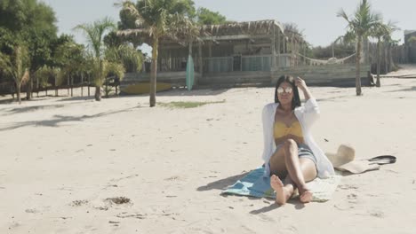 Feliz-Mujer-Hispana-Con-Gafas-De-Sol-Sentada-En-La-Playa-Bajo-El-Sol,-Espacio-Para-Copiar,-Cámara-Lenta