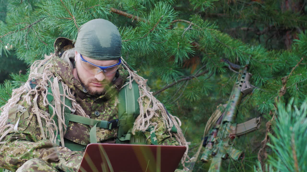 Portrett av en militærmann kledd i kamuflasje, sittende i skogen, som bruker en laptop.