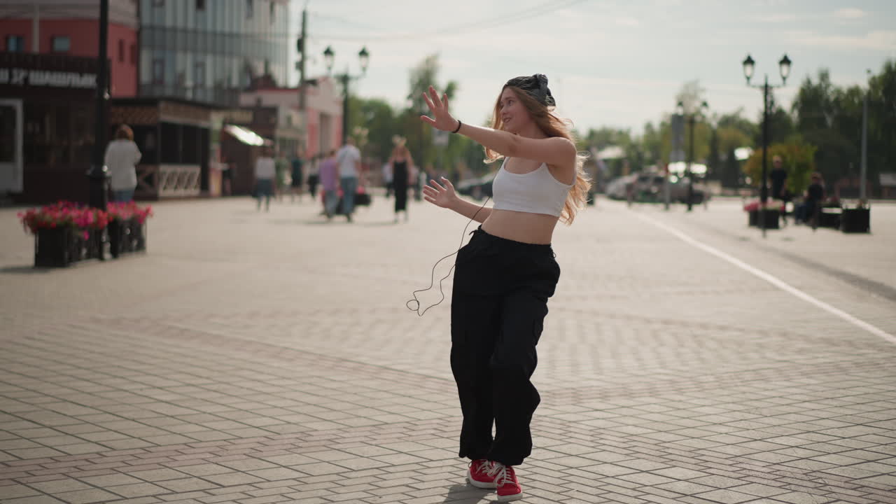 Žena běloška, pouliční tanečnice, předvádí v poledne na dlážděném náměstí optimistické freestyle pohyby, bílý crop top, černé cargo kalhoty, červené tenisky, bandana, expresivní gesta, kolemjdoucí
