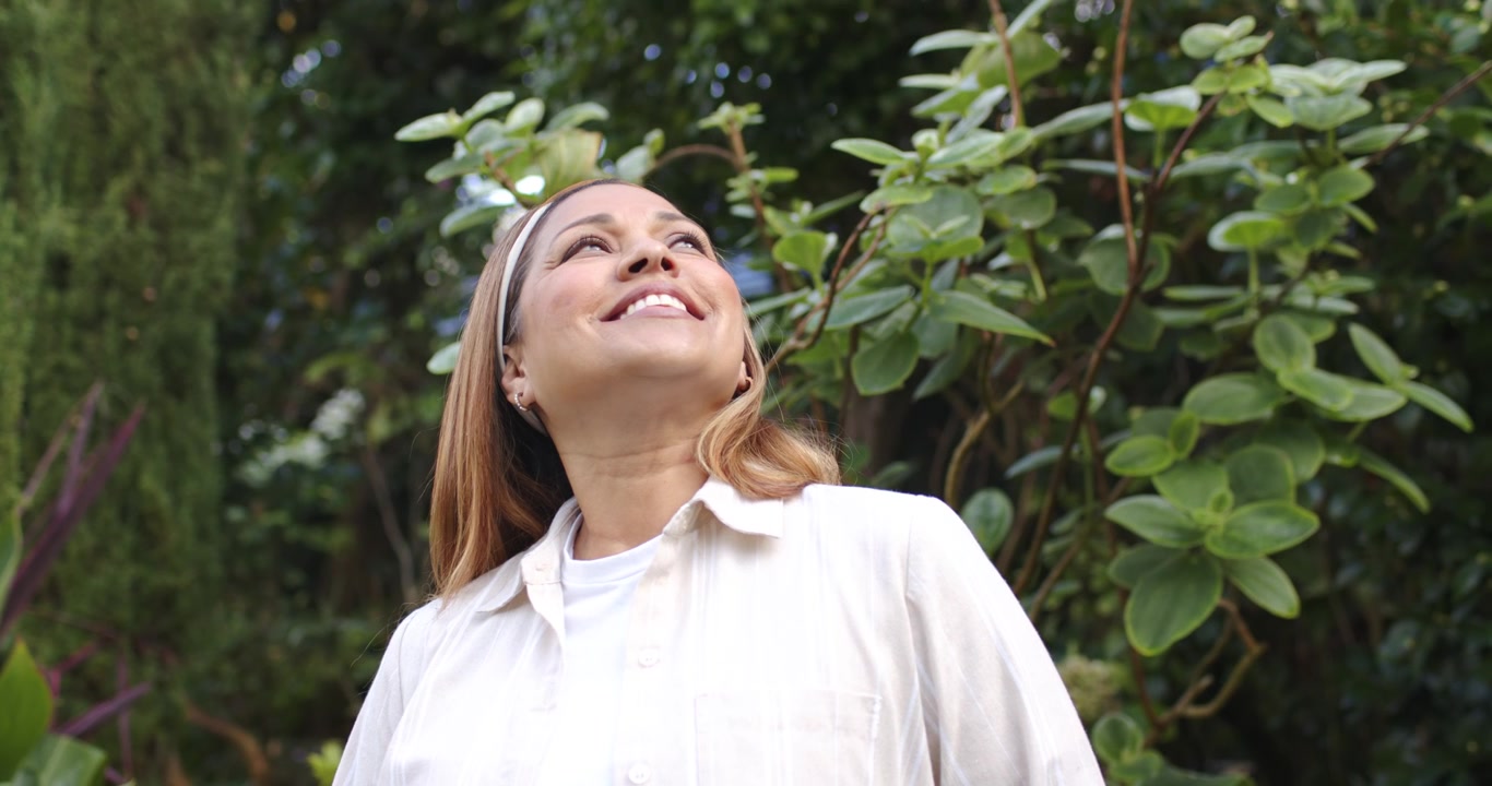 Donna afroamericana matura che vede un movimento in alto, si gira e sorride in giardino indossando una camicetta.