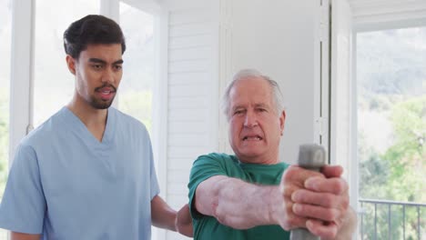 Biracial-male-therapist-talking-to-senior-man-exercising-with-dumbbells-at-retirement-home