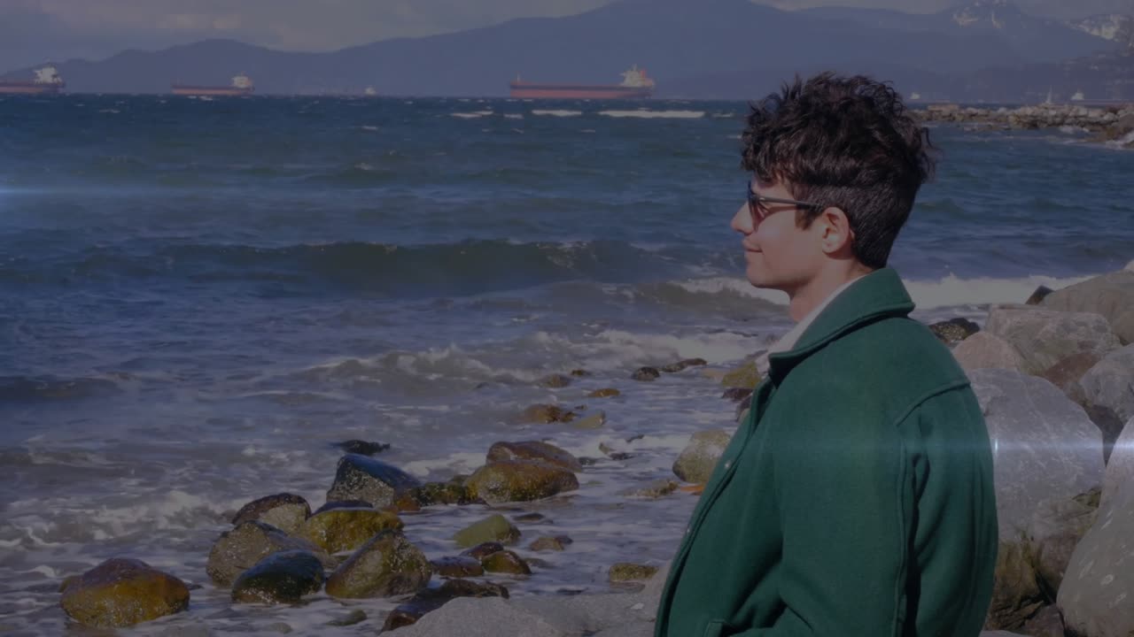 Uomo con cappotto verde che indossa occhiali da sole, con bagliori dell'obiettivo cinematografico che si propagano, rivelando una costa rocciosa, onde e navi.