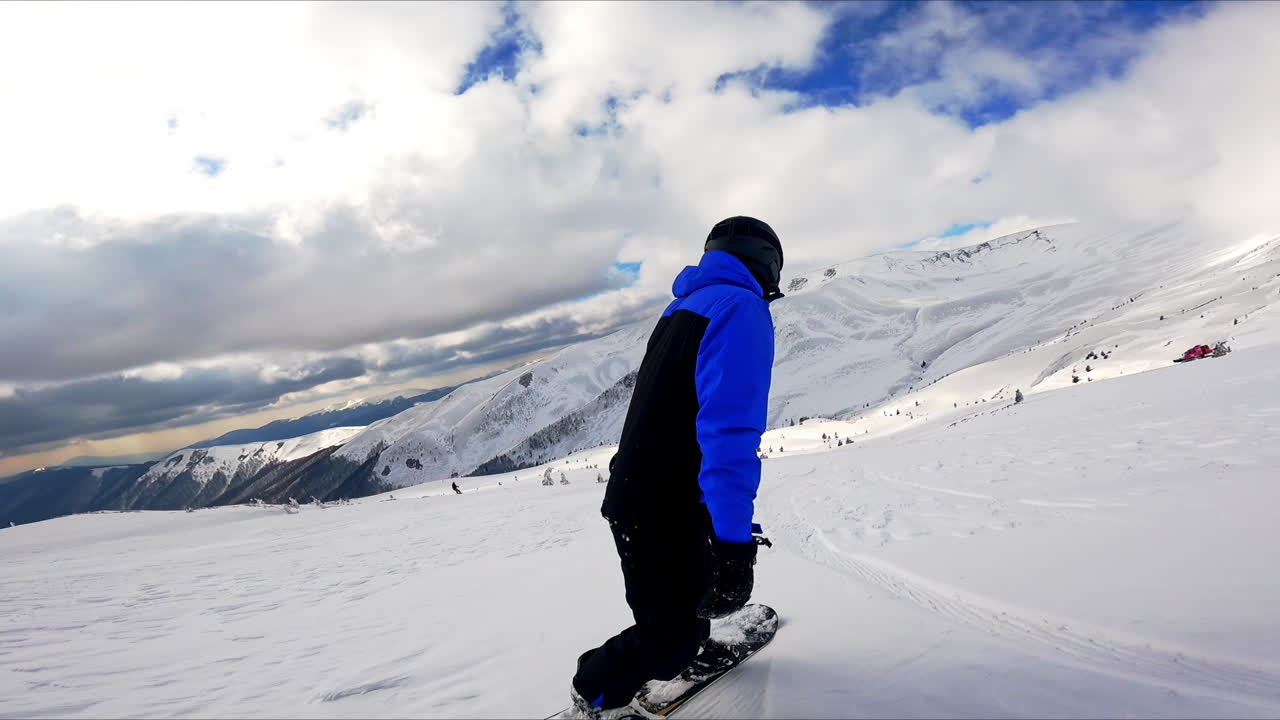 Professional snowboarder rides by the slope. Beautiful mountains under dramatic sky at backdrop.