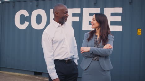 Portrait-Of-Male-And-Female-Freight-Haulage-Managers-Standing-By-Shipping-Container-For-Coffee