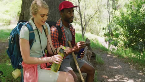 pareja feliz diversa con mochilas sentada y bebiendo agua en el parque, cámara lenta