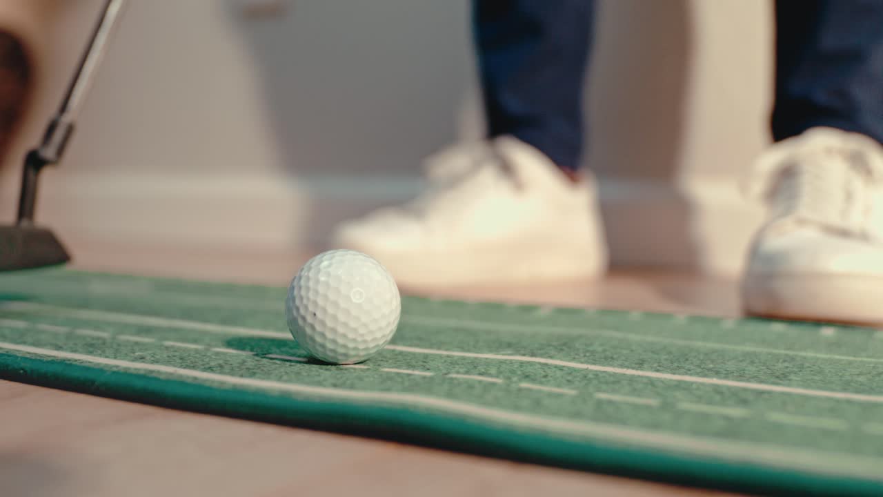 Primo piano al rallentatore mentre si posiziona una pallina da golf su un tappetino da allenamento per il putting indoor con scarpe da ginnastica bianche appoggiate e la faccia di un putter che si avvicina. Video di allenamento di golf e pubblicità di prodotti