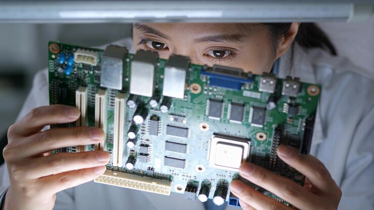 Raising PCB, adult Asian woman in white lab coat scanning board under lamp, verifying solder joints
