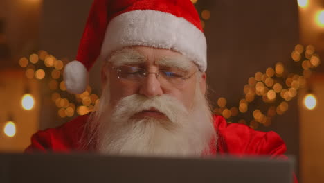 Real-Santa-Claus-using-new-technology-for-communication-with-children-receiving-mail-or-wish-list.-Cheerful-working-on-laptop-and-smiling-while-sitting-at-his-chair-with-fireplace-and-Christmas-Tree.-High-quality-4k-footage