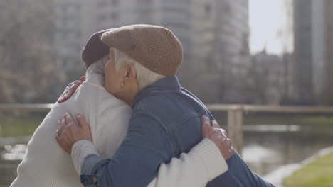 Affectionate-Elderly-Couple-Sitting-On-Park-Bench-In-Front-Of-Pond-And-Hugging