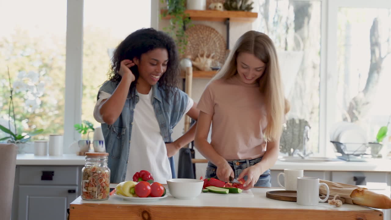 Two female friends, one juggles ingredients and the other cooks. Black girl and caucasian young woman talk and laugh in the kitchen. Medium shot.