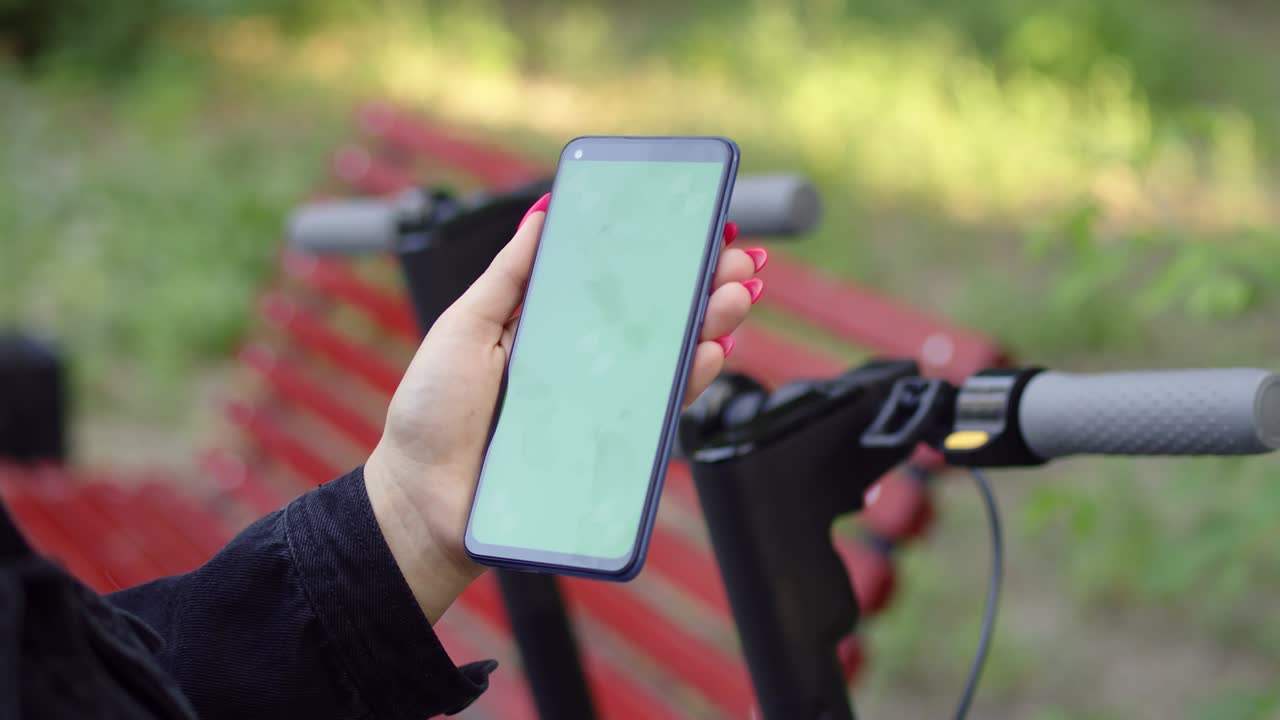 White woman's hand click phone with chromakey on background of electric scooter in public park, mockup, chromakey. Woman uses her phone on walk. Concept of leisure outdoor
