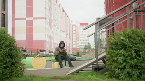 kid seated alone on outdoor bench with hooded jacket holding phone in hand, backpack beside, leaves swaying gently nearby, parked cars and tall residential buildings in background