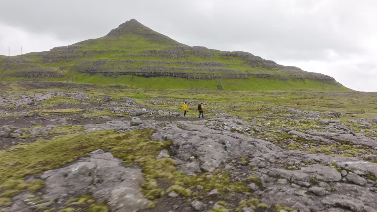 Two hikers walk in a rugged green landscape with rocky terrain, exuding adventure