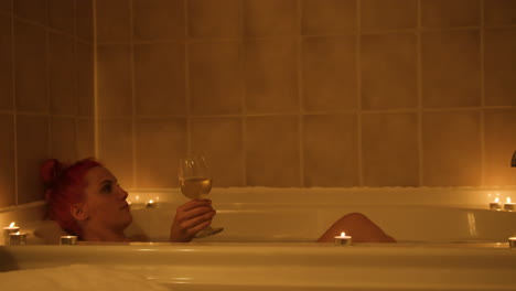 young woman reaches for glass of wine while taking a warm relaxing bath