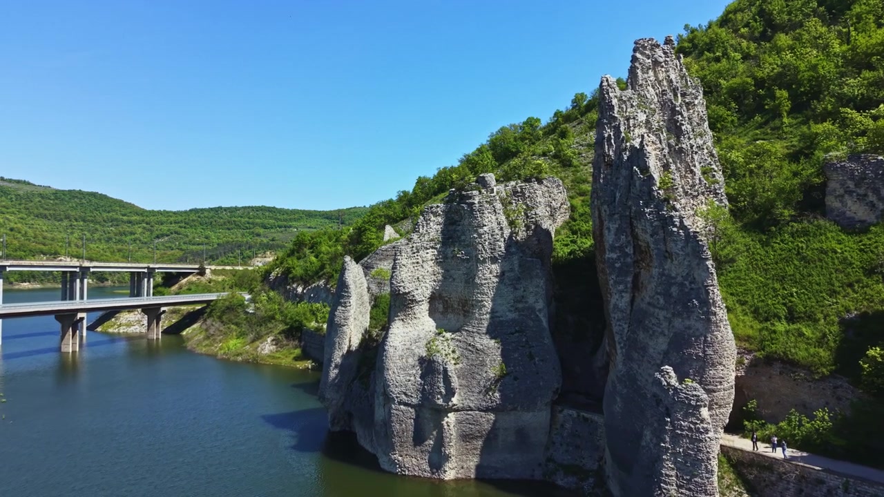 Rotsen steken boven een rivier uit in Bulgarije, bij een brug, met groene heuvels en een heldere hemel overdag