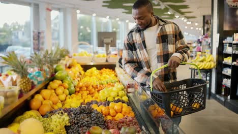 Un-Hombre-Moreno-De-Piel-Negra-Con-Una-Camisa-A-Cuadros-Y-Una-Camiseta-Blanca-Elige-Frutas-Cítricas-Y-Mandarinas-En-Un-Amplio-Mostrador-De-Una-Moderna-Tienda-De-Comestibles.