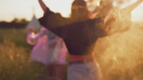 Two-Female-Friends-Camping-At-Music-Festival-Running-Through-Field-With-Smoke-Flares