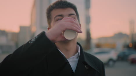 investor in black suit drinking coffee outdoors at dusk, glancing at something in background, background features building, parked cars, and modern structures. urban setting